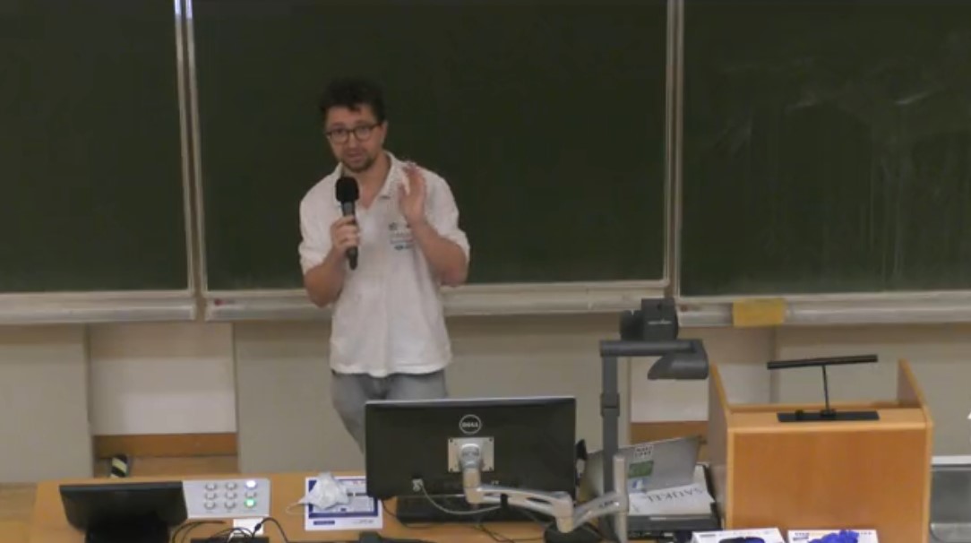 Screenshot der Vorlesung. Adrian steht mit Mikro vor der Tafel und erklärt etwas.